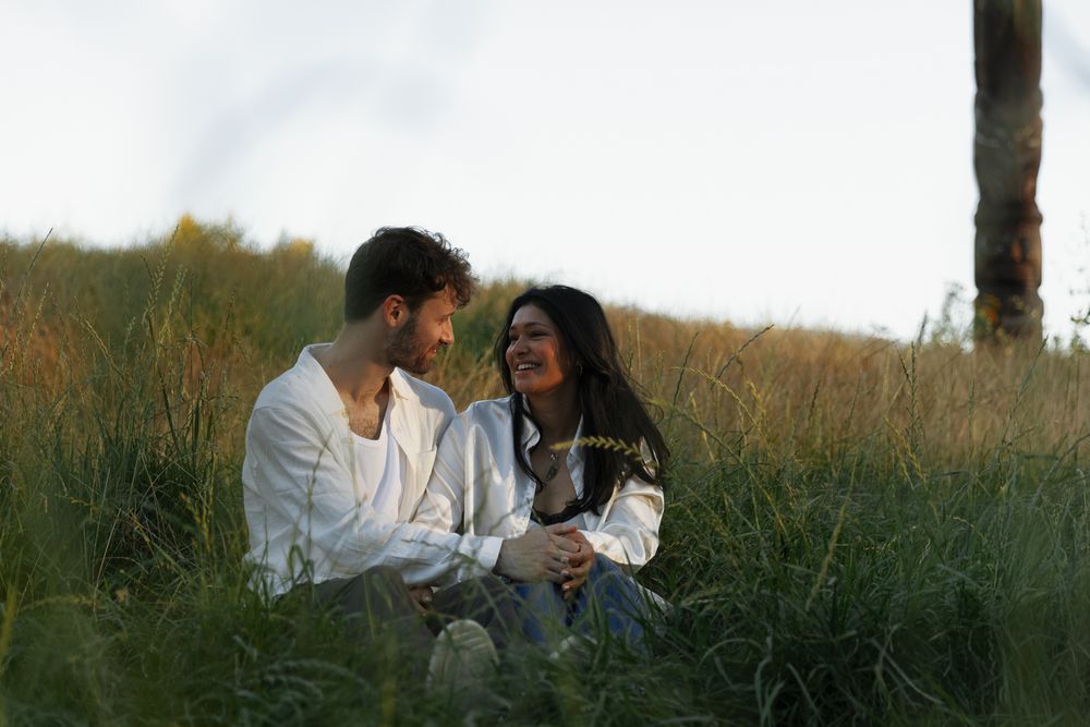 A couple looking lovingly at each other while sitting in some tall grass during a Vancouver engagement session taken by Kristina Harnett.