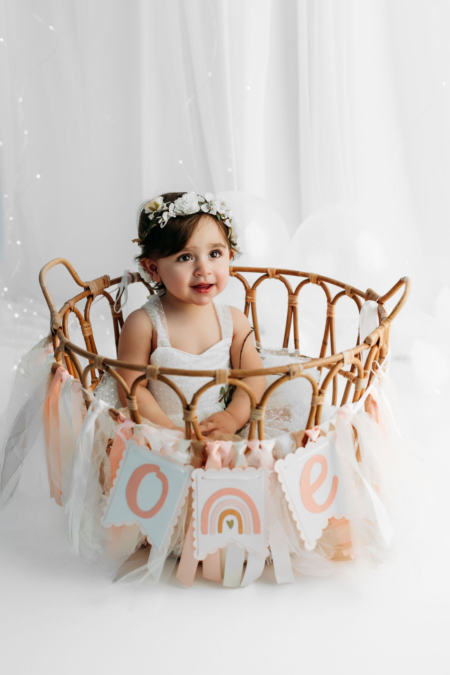 A baby in white sits on a light backdrop while posing with and eating a white birthday cake.