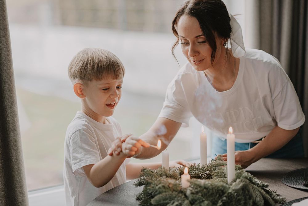 A parent and child share a special moment while lighting candles together at a table with greenery decorations.
