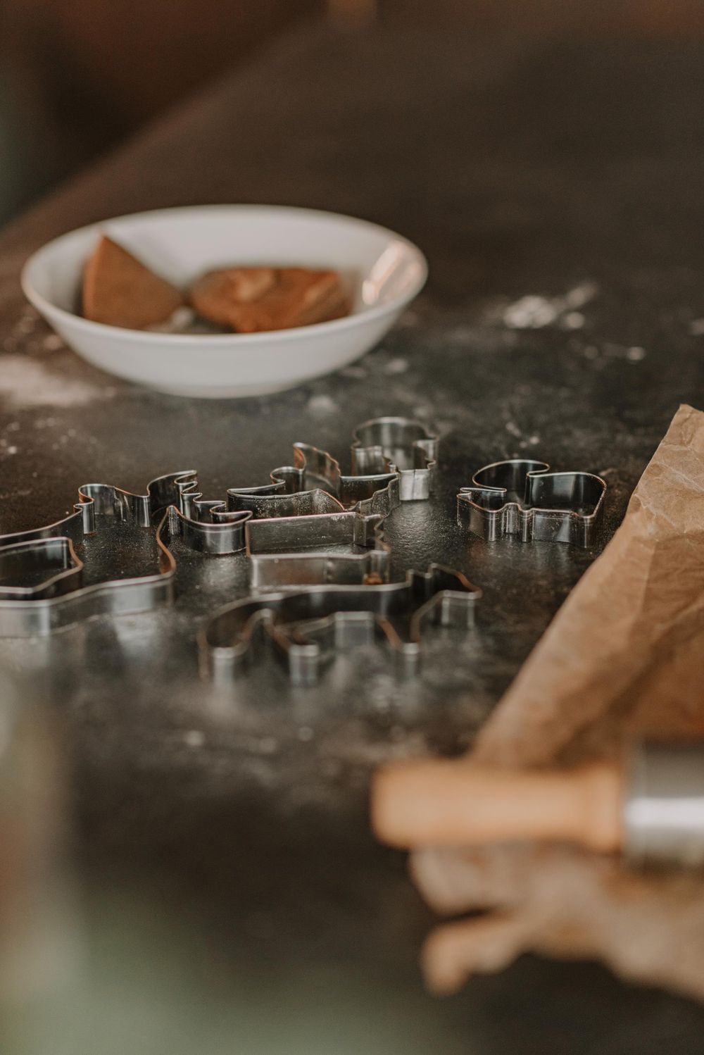 Metal cookie cutters lined up on a dark surface with brown paper and bowls in the background.