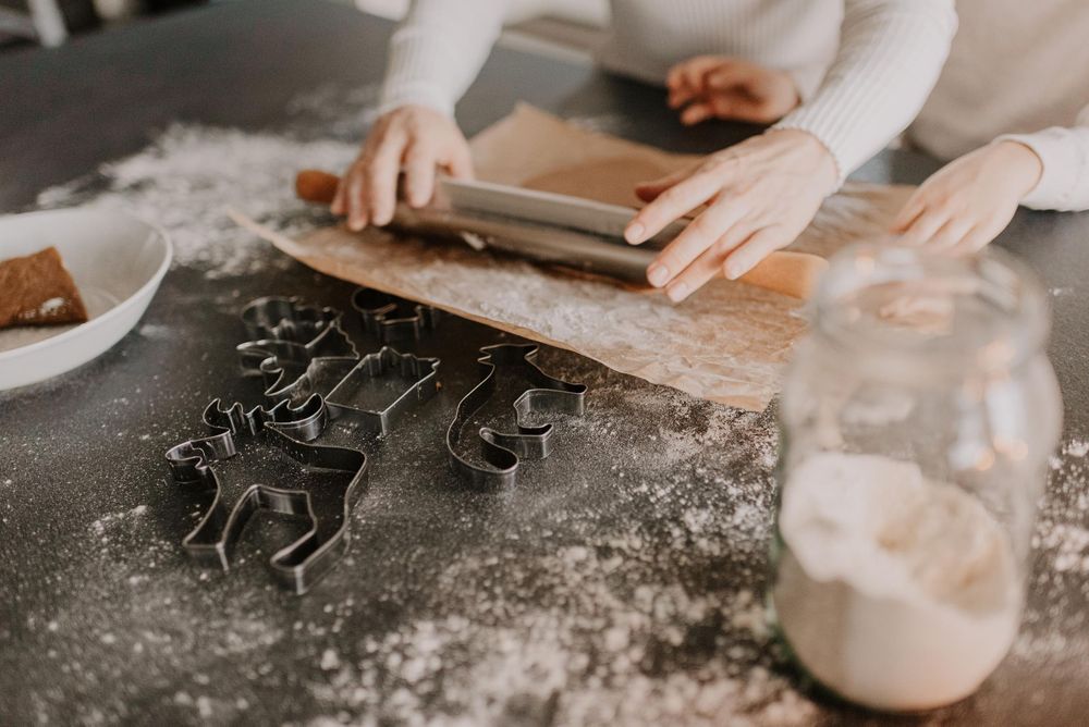 Hands rolling out cookie dough on a floured surface with cookie cutters and baking supplies nearby.