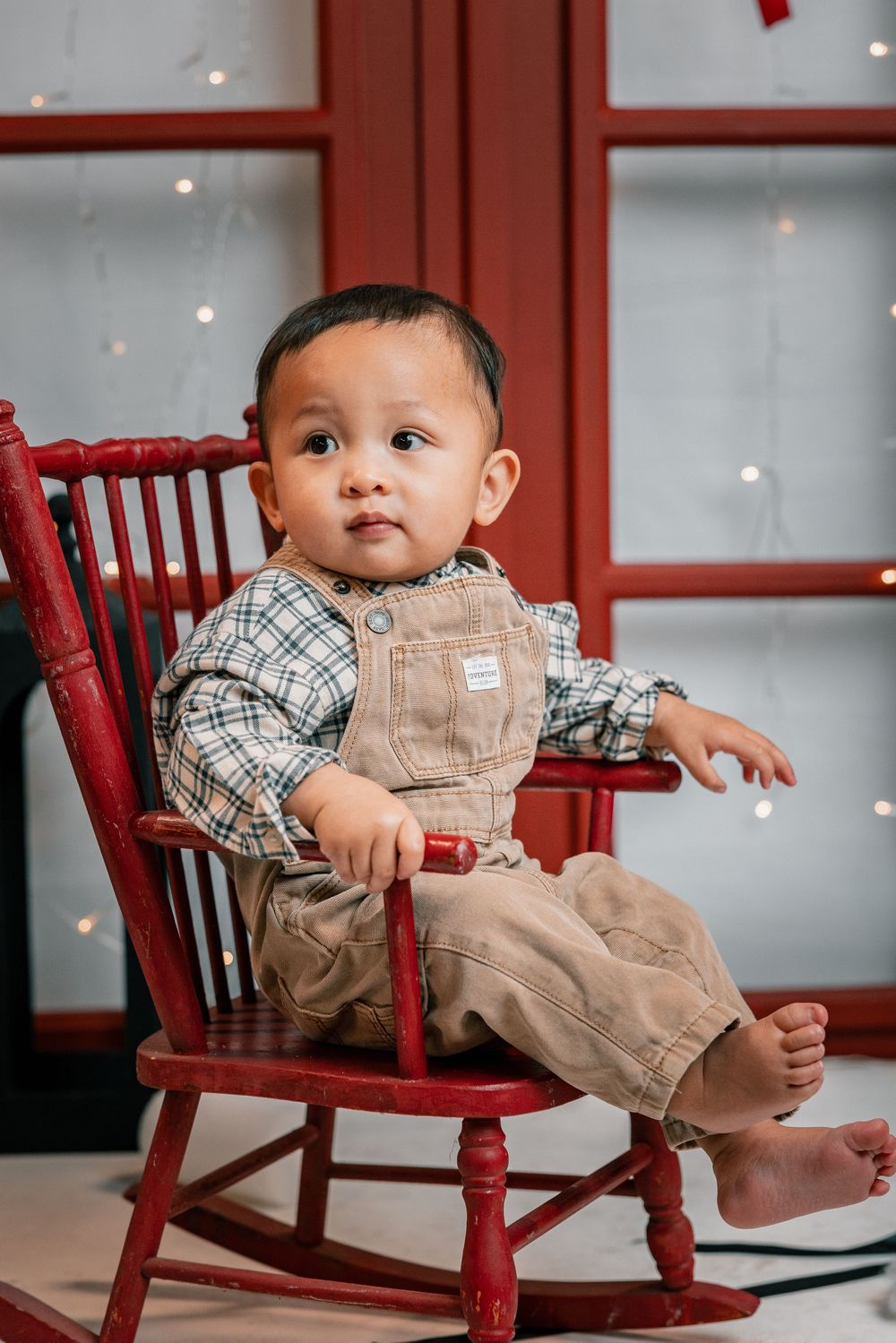 A baby in plaid shirt and tan overalls sits playfully on a red wooden chair with twinkling lights in the background.