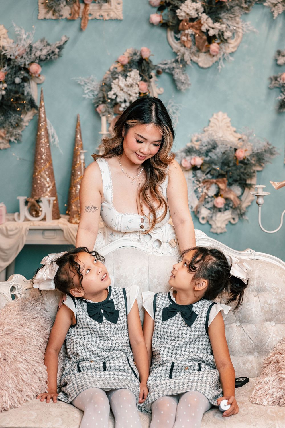 A woman in white dress sits with two young children wearing matching gingham dresses and bows against a floral backdrop.