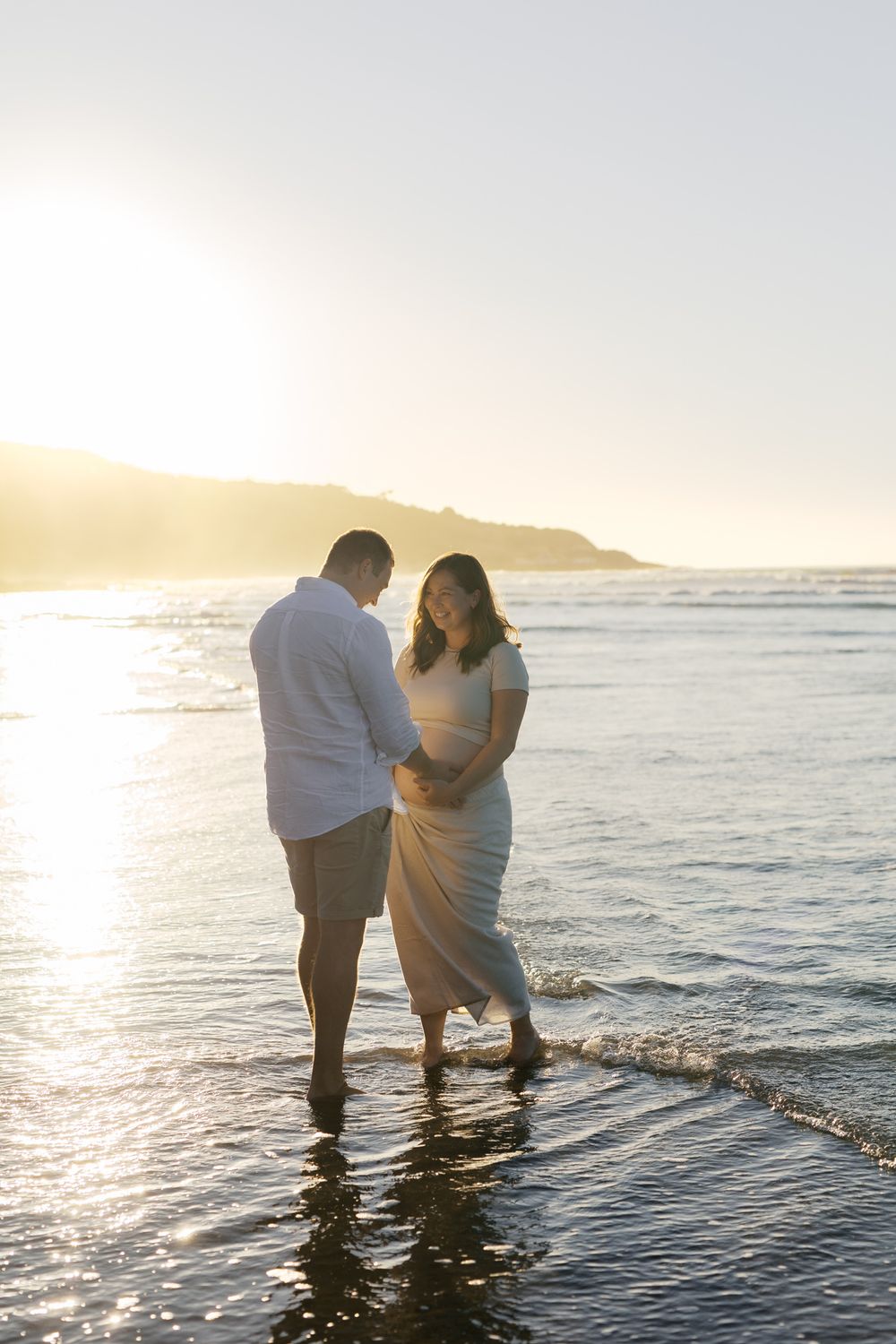 Two people stand together in shallow ocean water during golden hour sunset at the beach.