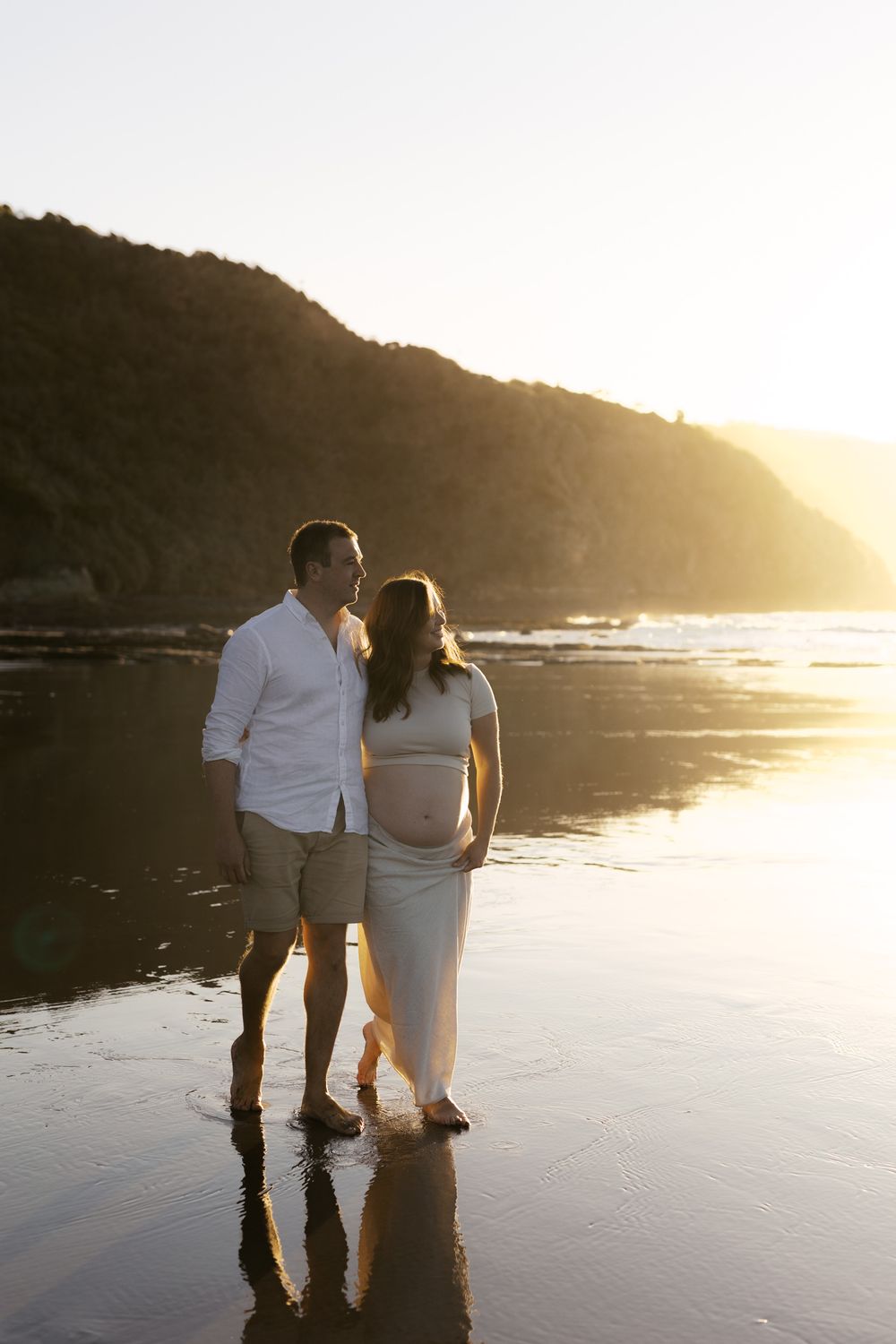 A couple walks together on a wet sandy beach at golden hour with cliffs and ocean waves in the background.