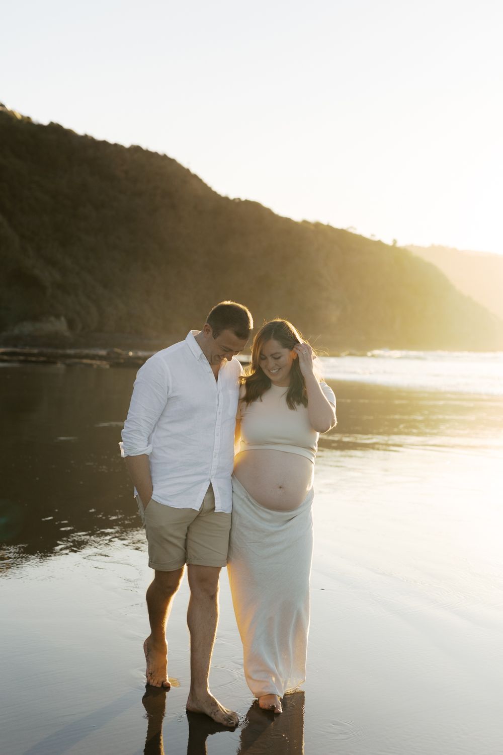 A couple shares an intimate moment on a beach at sunset with mountains in the background.