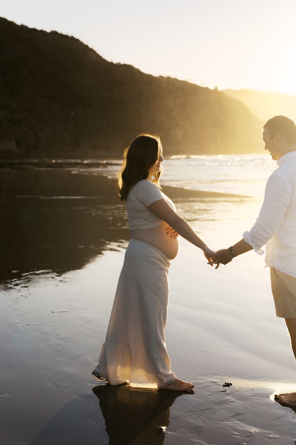 Couple in white clothing holding hands on a reflective beach at sunset.