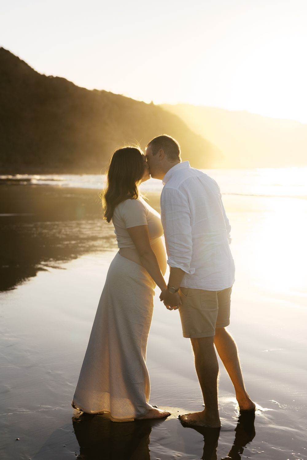 A couple shares an intimate moment on a beach at sunset with golden light reflecting off the water.