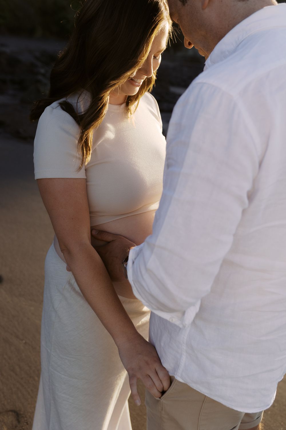 A couple holds hands intimately at sunset, wearing white and cream colored outfits on a beach.