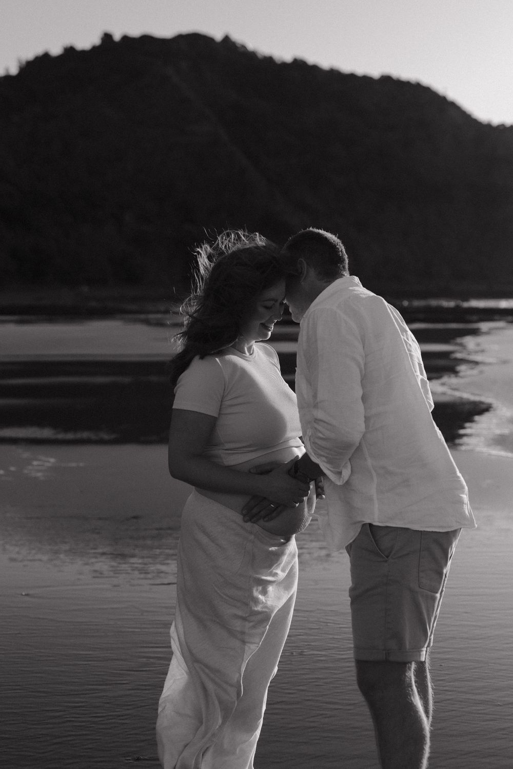 A romantic maternity photoshoot silhouette at sunset on a beach with mountains in the background.