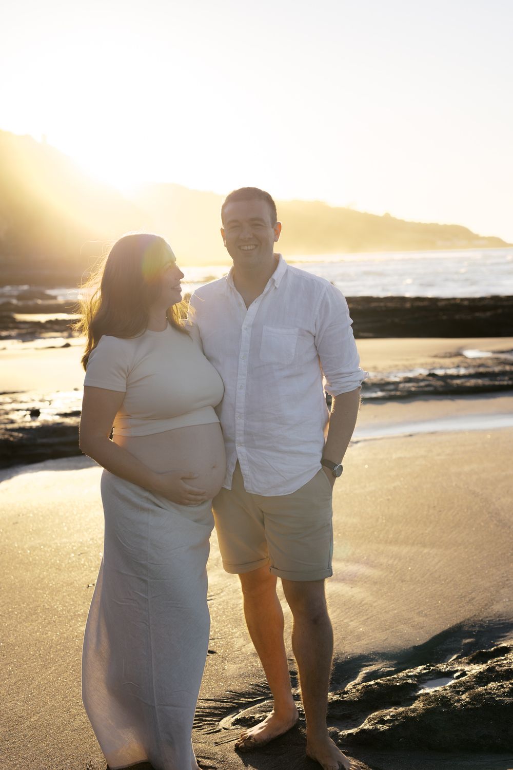 Two people in white clothing pose together on a sandy beach during golden hour.