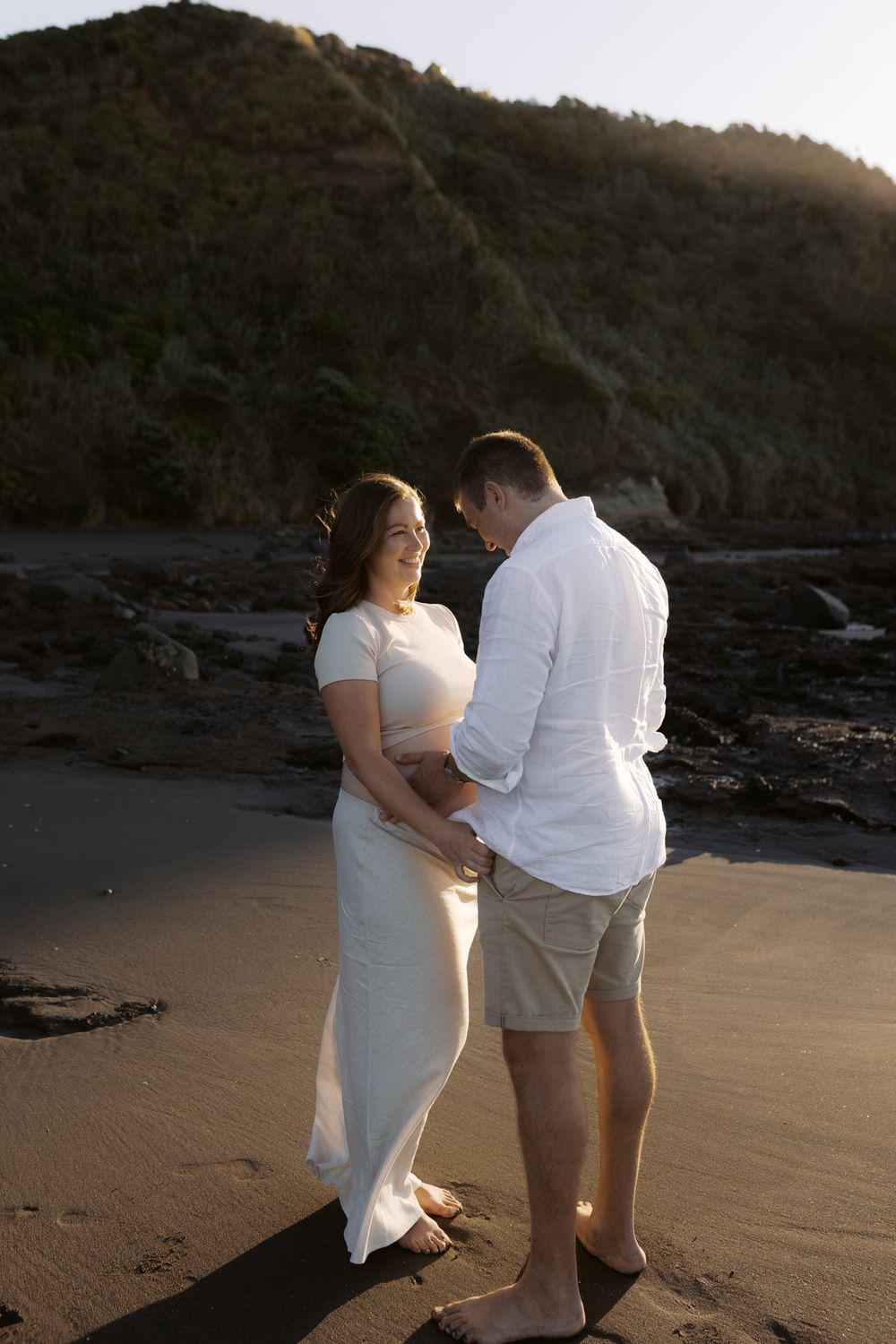 A couple in white attire shares an intimate moment on a beach at sunset with rocky cliffs in the background.