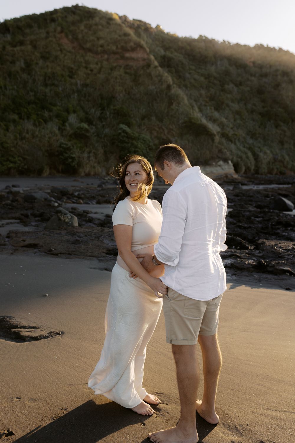 A couple shares an intimate moment on a rocky beach at sunset with mountains in the background.