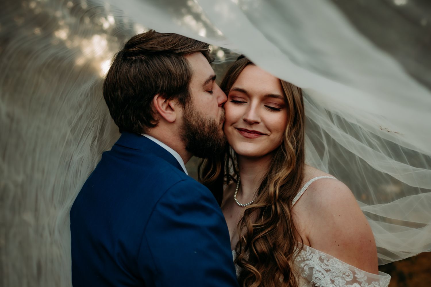 Intimate moment between newlyweds under flowing veil during outdoor portrait session.