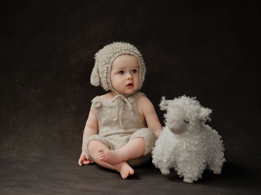 Baby in vintage-style cream knit bonnet and romper sits beside white plush teddy bear against dark background.