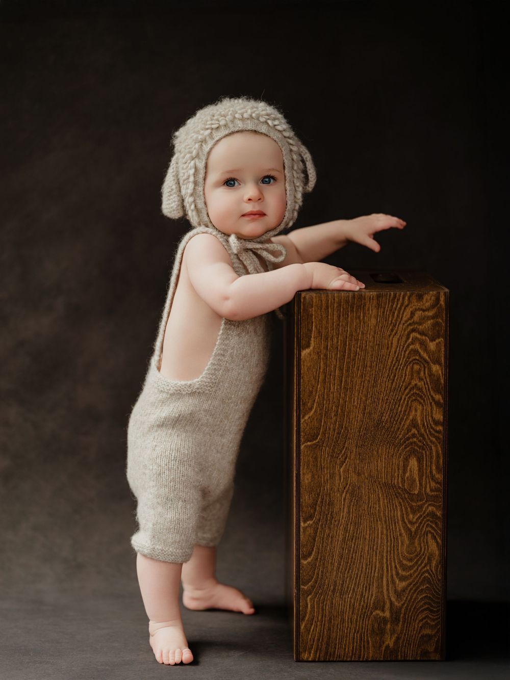 A small child in a knit bonnet and romper leans against a wooden box in a studio setting.