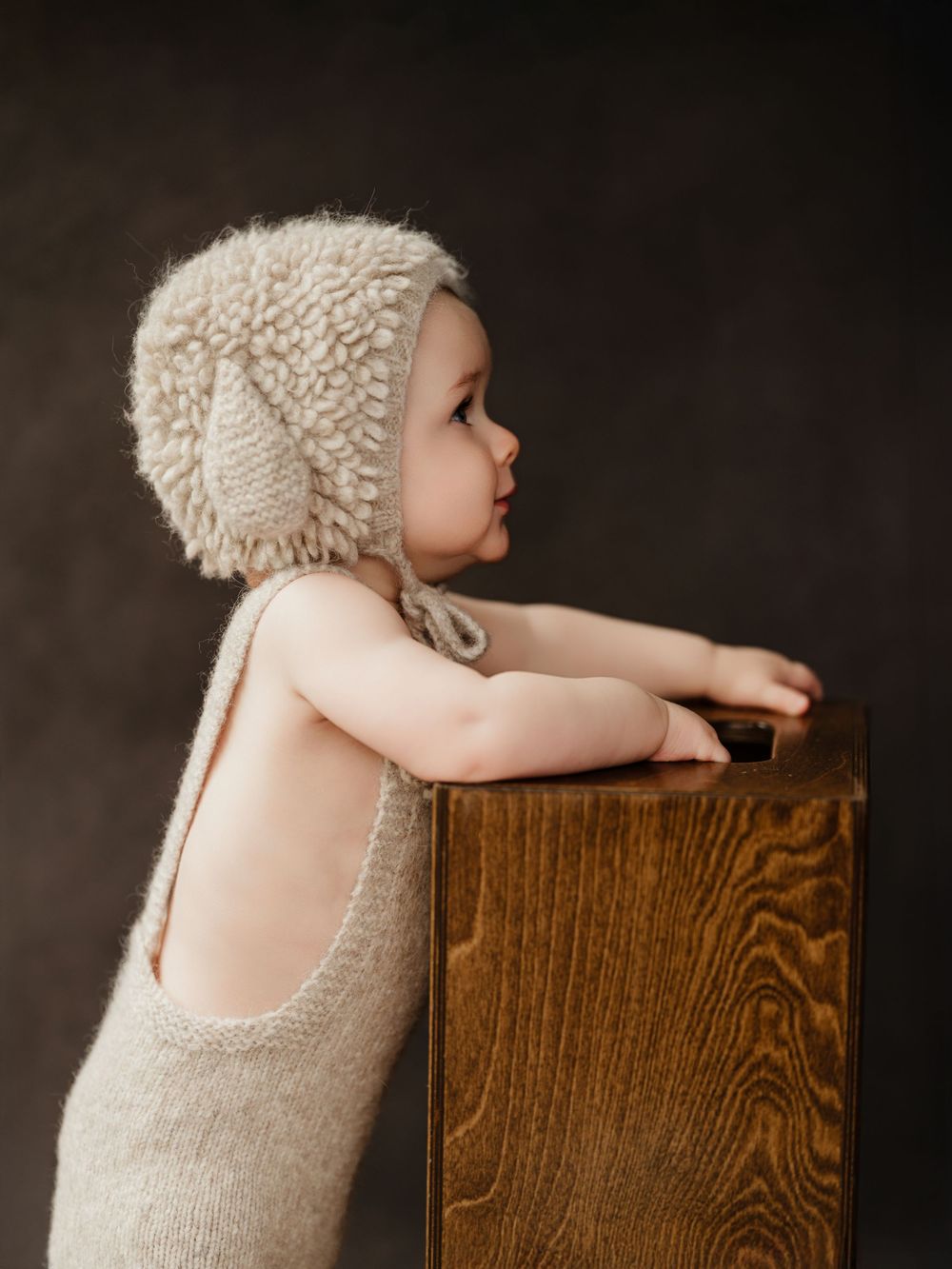 A baby wearing a knit bonnet and romper reaches up to touch a wooden surface in a studio setting.