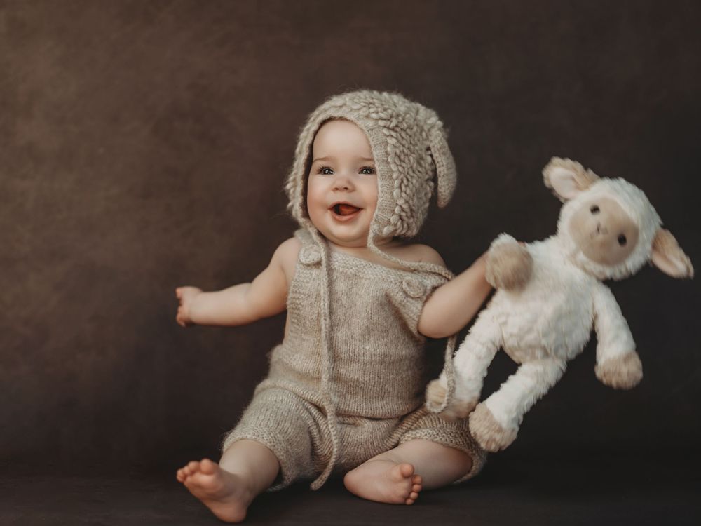 Smiling baby in knit outfit and bonnet hat sitting with a white lamb stuffed animal toy on brown background.