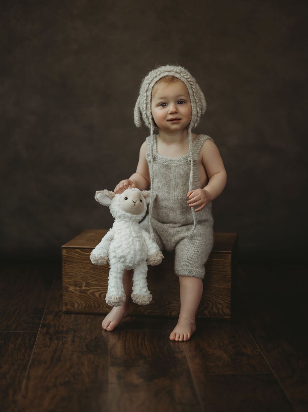Adorable toddler in knit lamb bonnet and linen romper sits on wooden box holding a fluffy white stuffed animal lamb.