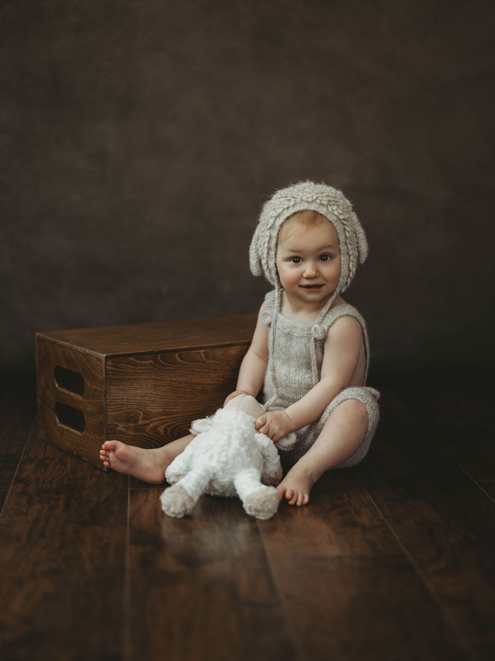 Smiling baby in knit bonnet and romper sits on wood floor with stuffed animal beside a wooden crate.