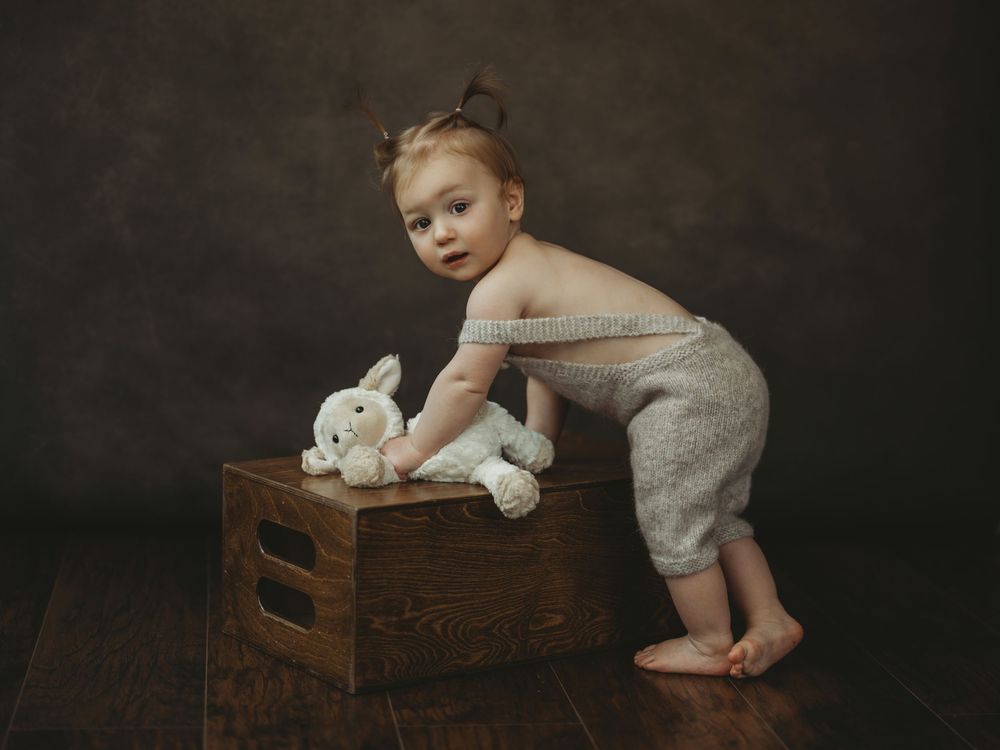 Toddler in gray romper leaning on wooden crate with white stuffed bunny, studio portrait on dark background.