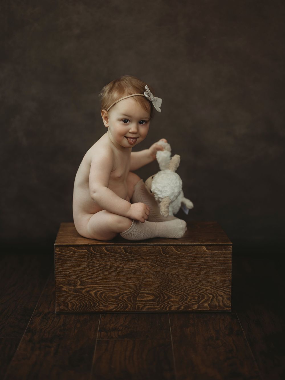 Adorable baby girl with bow headband sits on wooden box holding a white stuffed bunny toy.
