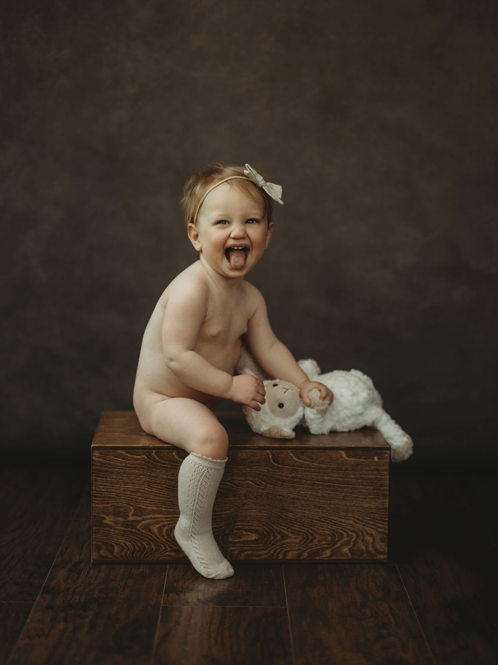 Laughing baby girl in knee socks and bow sits on wooden box holding a stuffed lamb toy against a dark studio backdrop.