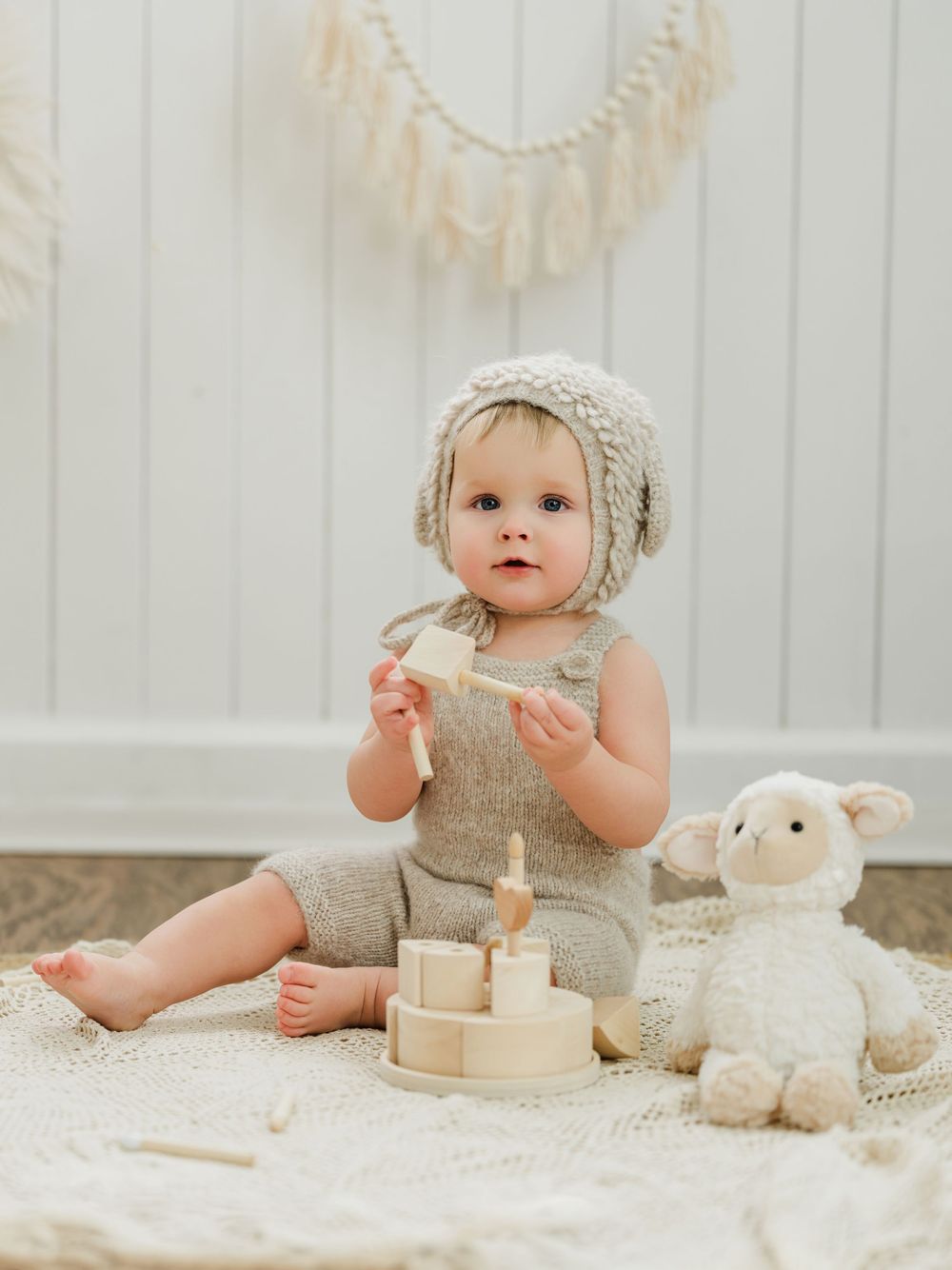 Adorable baby in knit outfit and bonnet playing with wooden stacking toy beside a plush lamb on a woven rug.