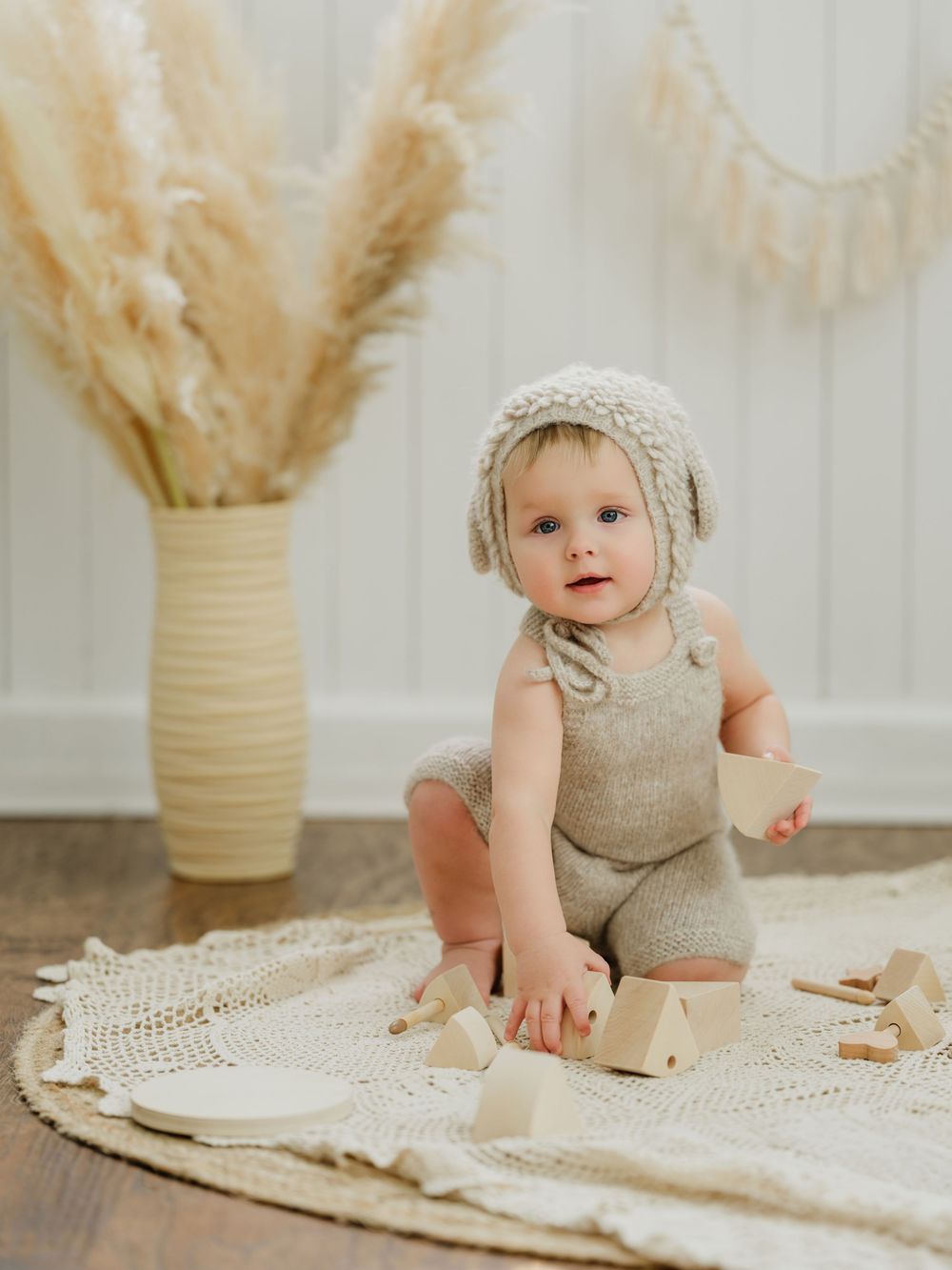 A cute baby in a knit bonnet and romper sits on a crochet rug playing with wooden blocks near pampas grass.