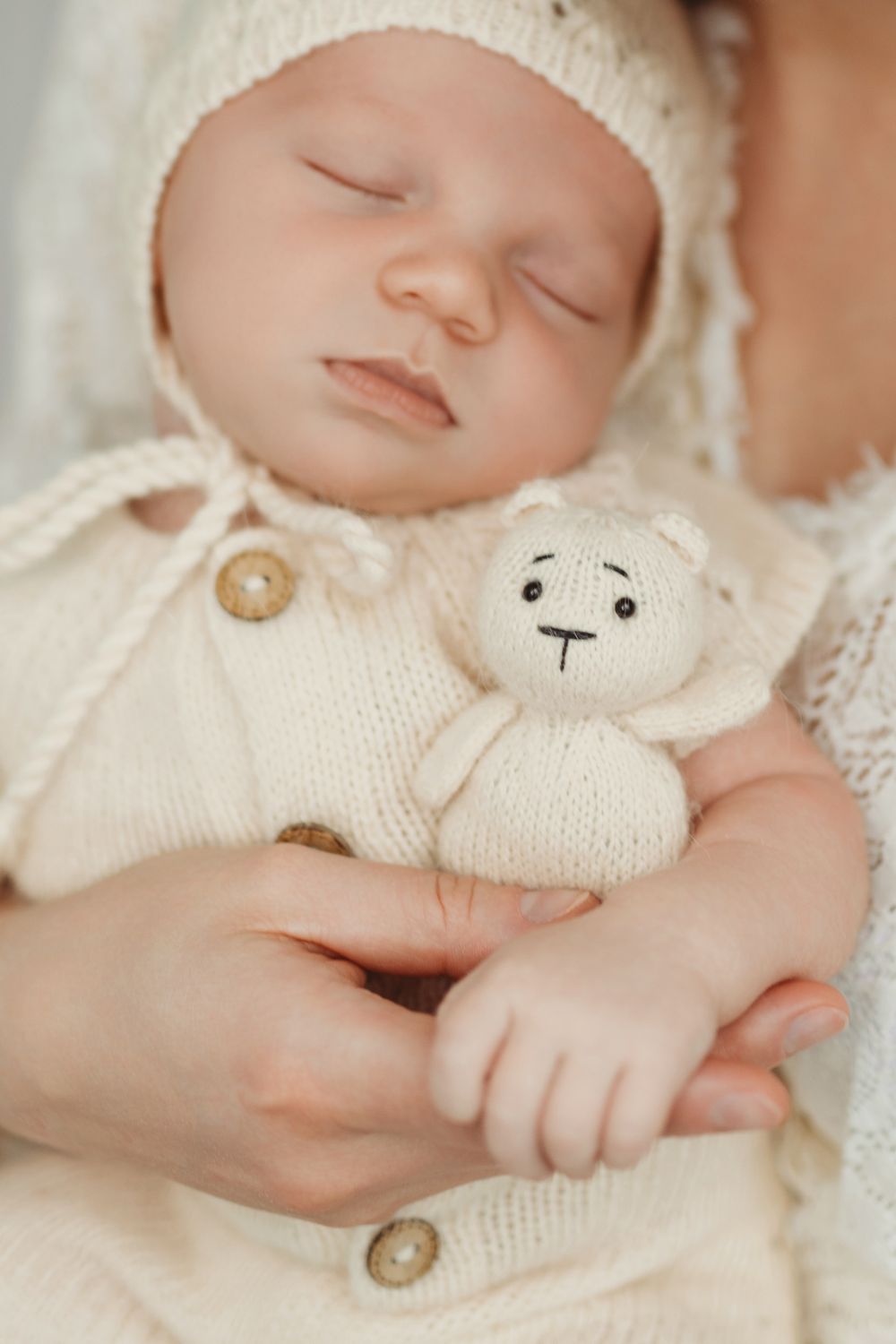 Peaceful sleeping baby in white knit clothing with small plush toy companion rests on soft cream-colored bedding.