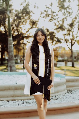 Person in graduation attire poses near fountain in outdoor setting at sunset.