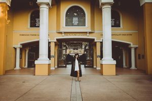Graduate poses in front of yellow Mediterranean-style building entrance with white columns.