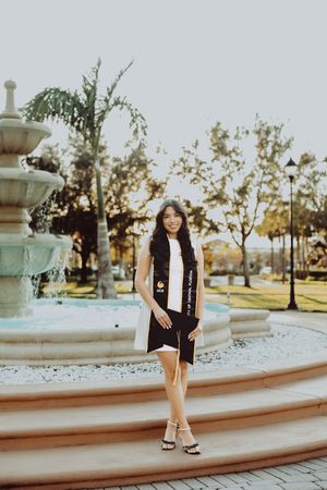 Graduate in black academic stole poses by curved fountain during golden hour.
