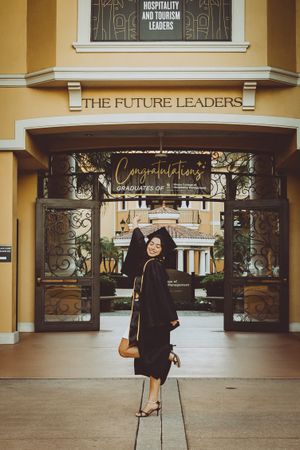 Graduate in black cap and gown stands in front of yellow building with hospitality signage.