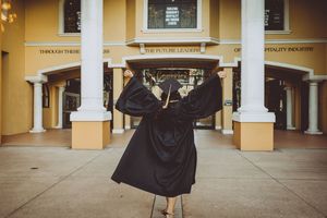 Graduate raises arms in celebration pose wearing black graduation gown outside yellow building.