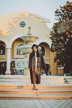 Graduate poses near decorative fountain outside Mediterranean style building at golden hour.
