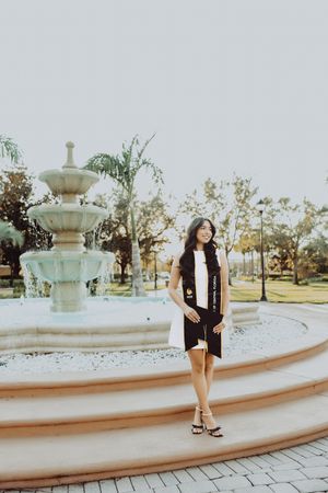 Graduate in black dress poses elegantly by ornate fountain in park during golden hour sunset.