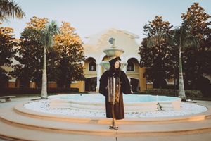 Graduate in black cap and gown poses in front of a white Spanish colonial building with a circular fountain.