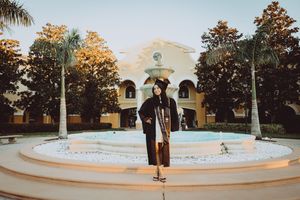 A panoramic view of a curved white fountain and Spanish colonial architecture at sunset with palm trees in the background.