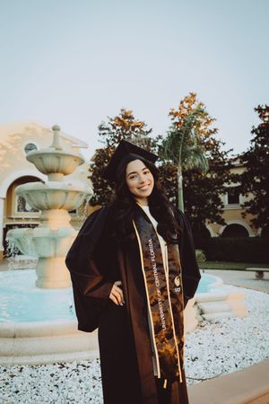Graduate in black cap and gown poses with a trophy by a decorative fountain at sunset.
