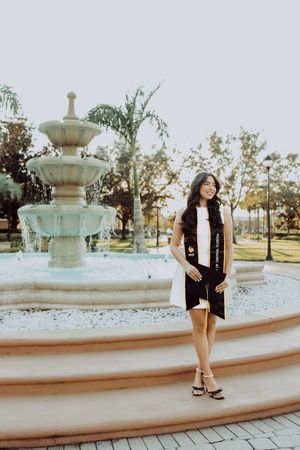 Graduate poses by ornate tiered fountain in outdoor campus setting at sunset.