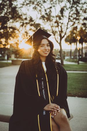 Graduate in cap and gown poses at sunset with trees in background.