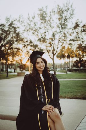 Graduate in black cap and gown sits outdoors during golden hour.