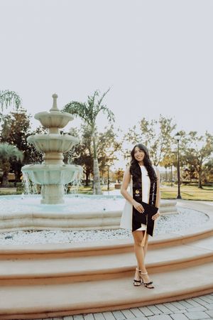 Graduate in white dress and stole standing by ornate fountain during golden hour.