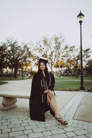 Graduate in black gown sits on stone bench near lamp post during golden hour.