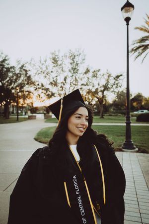 Graduate in black cap and gown stands by lamppost at sunset on campus walkway.