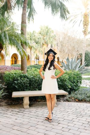 Graduate in white dress poses outdoors with palm trees and stone pathway in tropical garden setting.
