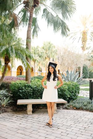 Graduate in white dress walks along tropical palm-lined pathway.