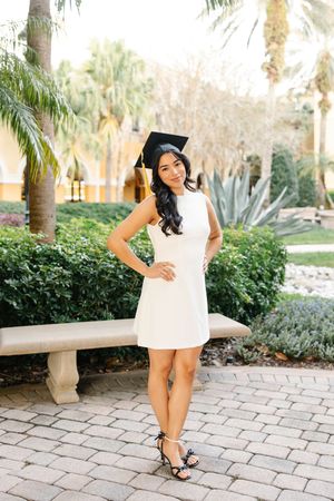 Graduate in white dress poses near palm trees and landscaping on a brick paved path.