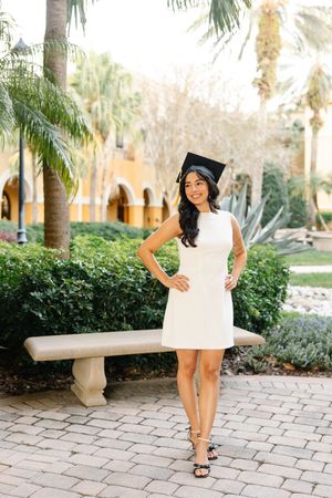 Graduate in cap and gown poses on brick pathway with palm trees.